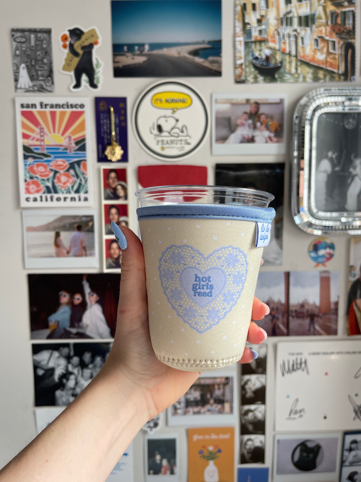 A hand holding a clear plastic travel tumbler with a light blue fabric sleeve. The sleeve features a light blue heart pattern with the text 'hot girls read' in the center.